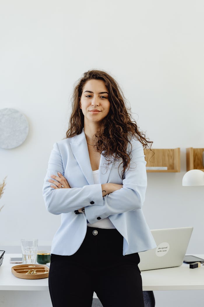 Professional woman in a white blazer standing confidently in an office with arms crossed.